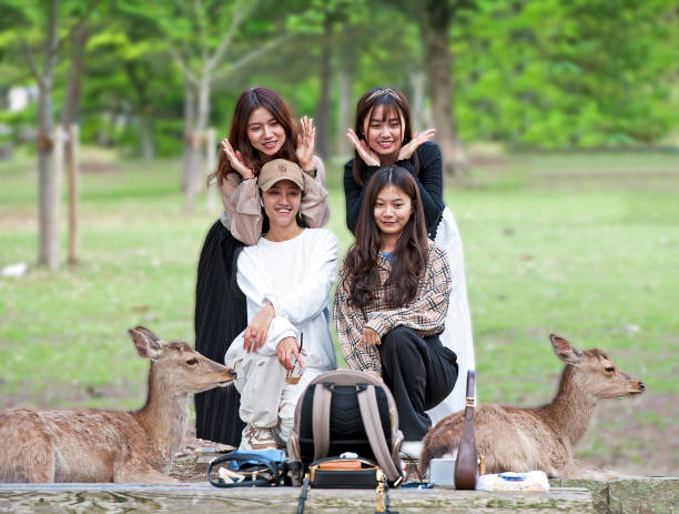 Young women pose for a selfie amidst deer, Nara Park, Nara, Nara Prefecture, Japan. Nara once capital of Japan is now the capital of Nara Prefecture, with significant temples and artwork dating to the 8th century. Deer roam in Nara Park, site of Tōdai-ji temple. Daibutsu. Honshu, Japan’s main island with most of the country’s major cities and cultural sites. From the busy global hub of Tokyo for finance, industry, fashion and cuisine to Mount Fuji, the iconic, snow-capped volcano and the city of Kyoto, famed for Zen temples, gardens and traditional geisha entertainment