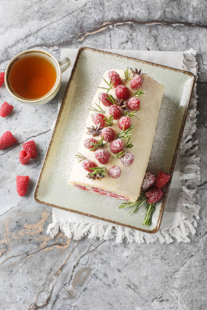 istock Festive raspberry sponge roll cake with jam, berries, cream cheese covered with white chocolate close-up in a plate on the table. Vertical top view 2157318655