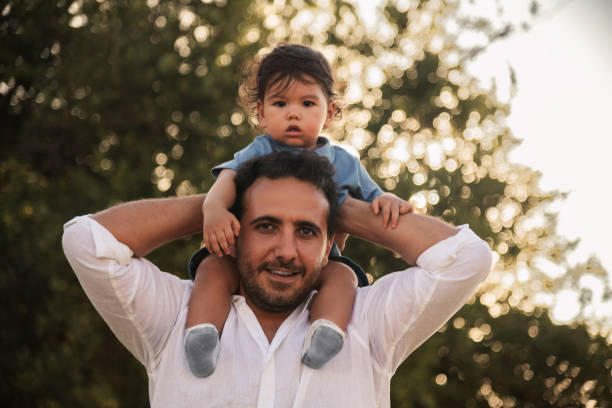 A one-year-old boy sitting on his father's shoulders with trees and bokeh in the background, capturing a special moment on Father's Day