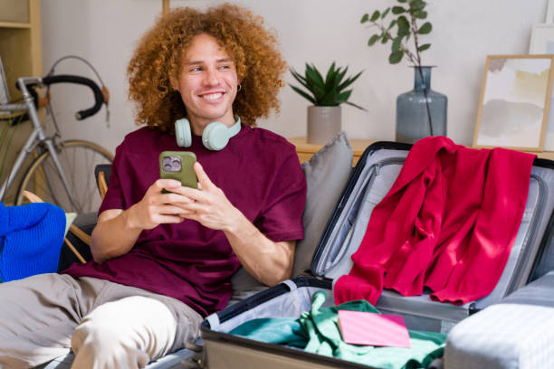 istock Young man sitting on sofa with packed suitcase browsing on smartphone 2157349348