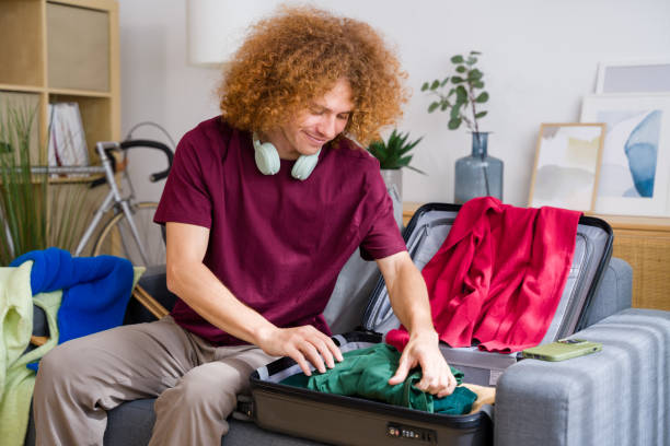 istock Happy young man packing clothes for trip sitting on sofa at home 2157349394