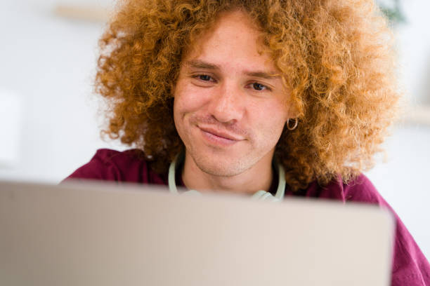 Young man with curly red hair smiles while working from home on his laptop, showing contentment and productivity