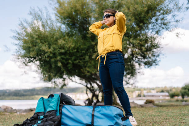A woman in a yellow jacket stands in front of a tree with a backpack and a blue bag