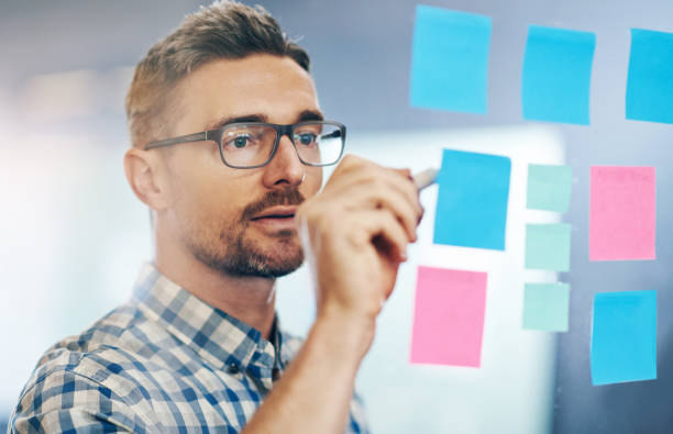istock Man, writing and sticky notes in office for planning, tasks and schedule with glasses. Businessperson, strategy and idea on paper for calendar, brainstorming and agenda on glass wall in workplace 2157354709