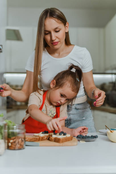 istock Mother Watching Small Daughter Put Bananas And Berries On Peanut Butter Bread 2157356819