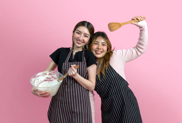 Two women are having fun baking together. They are wearing aprons and smiling while mixing ingredients with a wooden spoon. Portrait on pink background with studio light.