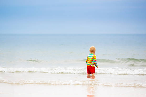 Kids playing on tropical beach. Children swim and play at sea on summer family vacation. Sand and water fun, sun protection for young child. Little baby running and jumping at ocean shore.