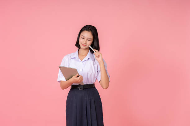 istock Asian female high school student, presenter and looking at laptop, using pen, thinking about education, studying online in school system, taking photo in studio, pink background 2157392271