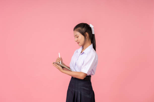 istock Asian female student with long hair Side view using laptop to work, submit to teacher in eclipse, study online for final exam, wear school uniform, take photo in studio, pink background 2157392299
