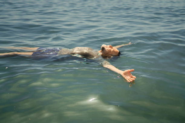 The guy carefully leads the girl out of the water onto the shore after she was covered by a wave. She walks with her eyes closed against the seawater that has entered them.