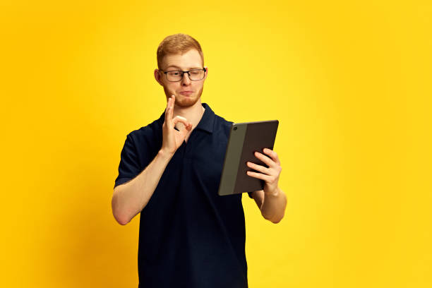 istock Approval. Young man in glasses looking on tablet with evaluating face, checking and approving information against bright yellow background 2157396427
