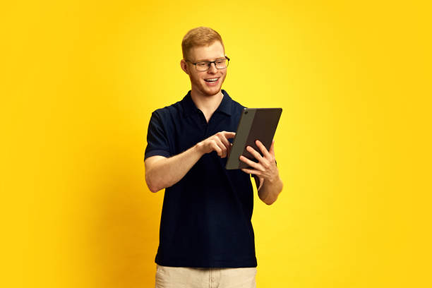 istock Smiling young redhead man in glasses looking at tablet and pointing with finger, using online services against bright yellow background 2157396448