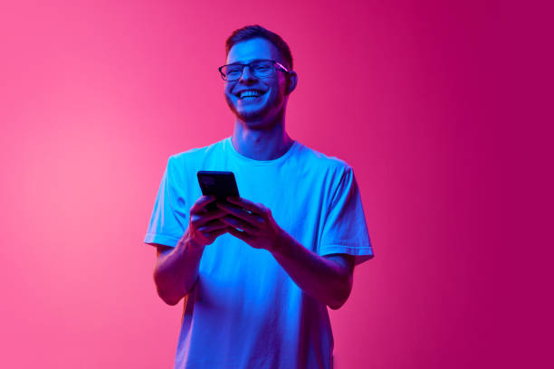 istock Laughing young man in glasses and casual clothes using mobile phone for communication against pink background in neon light 2157396472