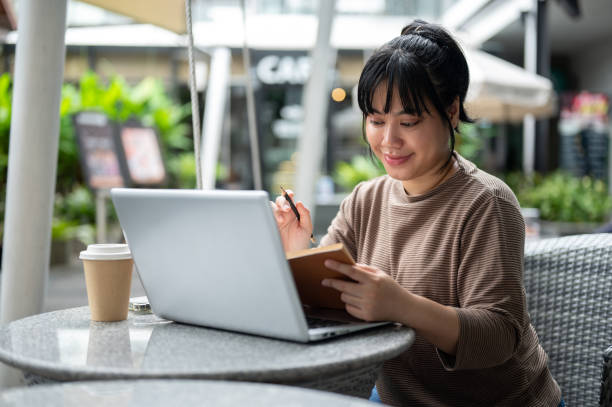 istock A woman is working remotely at a cafe, sitting outdoor, using her laptop and reading a book. 2157397833