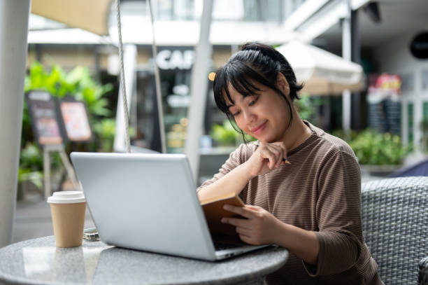 istock A woman is working remotely at a cafe, sitting outdoor, using her laptop and reading a book. 2157397835