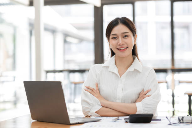 istock Confident Business Woman Working in Modern Office Environment with Laptop and Documents, Smiling and Looking at Camera, Professional Female Entrepreneur in White Shirt, Bright and Airy Workspace 2157418268