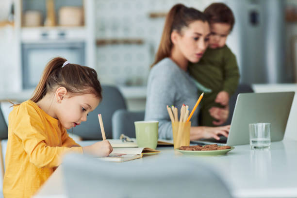 istock A creative small girl drawing in her notebook while her mother is working in the background and holding a toddler 2157421661