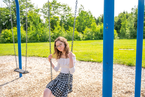 A woman with pigtails in youth denim outfit sits on a wooden swing in a park shady area.