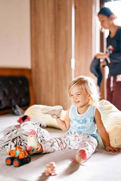 istock Little girl with a sneaker in her hand sits on a bed among toys 2157444773