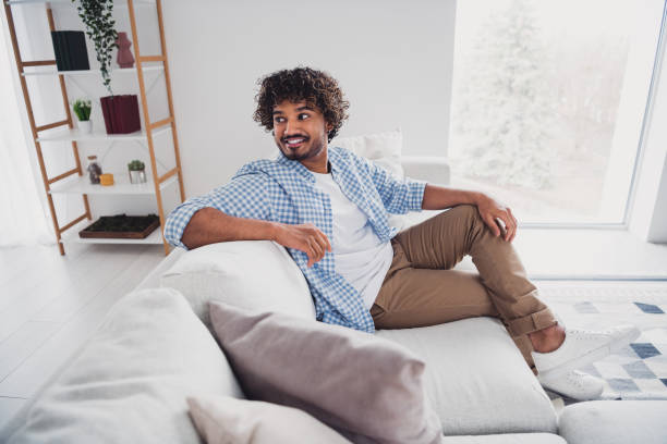 istock Photo of cheerful dreamy guy dressed plaid shirt smiling enjoying weekend sitting sofa indoors room home house 2157446033