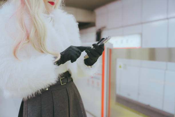 A woman wearing a white fur coat and black gloves is using her smartphone while waiting for the subway.