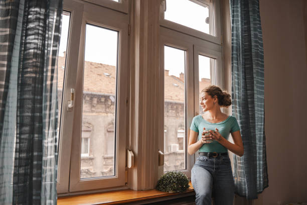 istock Young woman looking through the apartment window while drinking her coffee 2157453433