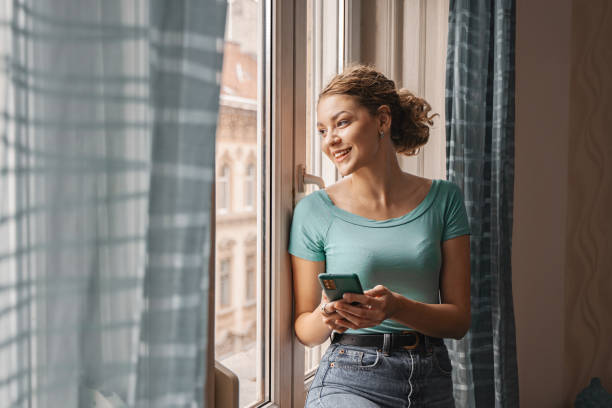 istock Young woman looking through the apartment window while using her mobile phone 2157453438