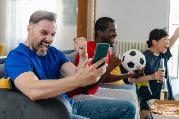 istock Cheerful mid adult man using smart phone while watching sports with friends 2157455607