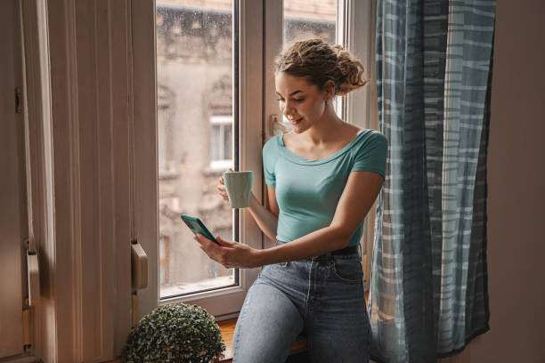 istock Young woman drinking coffee and using mobile phone beside apartment window 2157458669