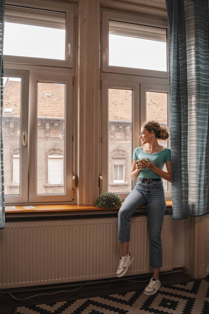 istock Young woman looking through the apartment window while drinking her coffee 2157459428