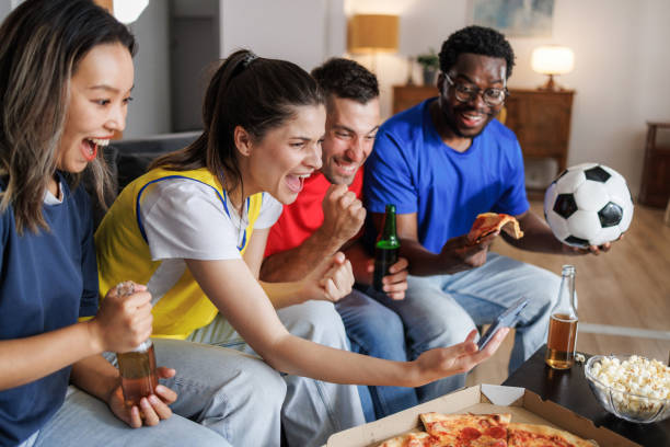 istock Multiethnic friends sharing pizza, popcorn and beer while watching a soccer game on a smart phone. 2157486524