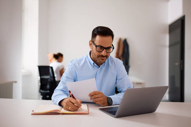 istock Smiling businessman writing notes while working on laptop in the office. 2157492214