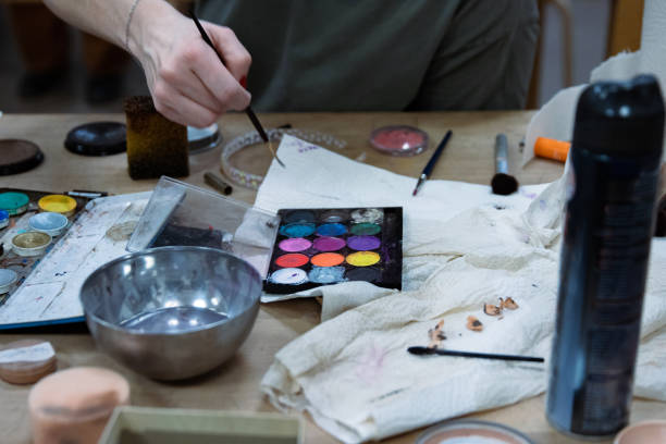 istock Messy colorful creative table with makeup palette, paints, brushes, pencil sharpener, paintbrushes set and bowl of water, paper tissues on wooden table. Stage make up. Creative chaos. Selective focus 2157494649
