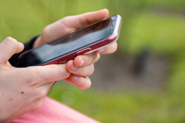 Thumb finger on black screen of red mobile phone. Person texting text message, hipster touch smartphone screen. Girl hands holding scrolling working. Summer park. Close up. Copy space. Selective focus
