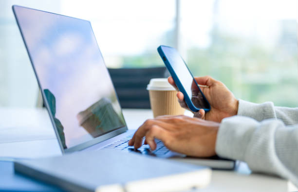 Close up of a Businessman working on a laptop computer and holding and looking at a mobile phone in the office. Close up of a Businessman working on a laptop computer and holding and looking at a mobile phone in the office. There are notepads on the desk. He is casually dressed text photography stock pictures, royalty-free photos & images