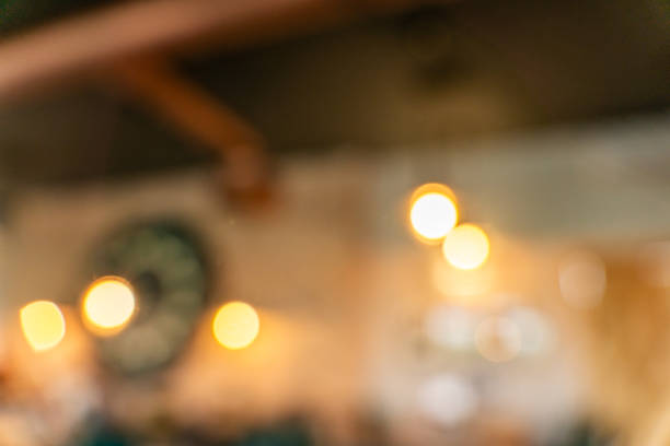 This image showcases a blurred, out-of-focus background of a cafe interior. The warm, inviting atmosphere is evident through the soft glow of hanging light fixtures, creating a bokeh effect. The image captures the essence of a cozy cafe, inviting the viewer to imagine the sights, sounds, and smells of the space.