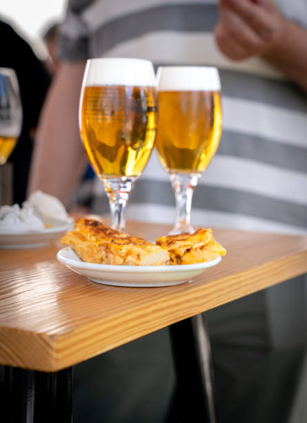 This image features two glasses of beer with frothy tops on a wooden table, accompanied by a plate of Spanish omelette (tortilla espanola) tapas. The scene captures a casual dining moment with the focus on the golden beers and the delicious tapas. The blurred background shows a person in a striped shirt, adding a relaxed and social atmosphere to the setting. This image is perfect for showcasing traditional Spanish cuisine and the conviviality of enjoying tapas with friends
