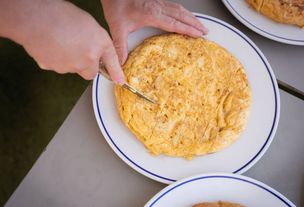 This top view image captures the moment of slicing a traditional Spanish omelette (tortilla espanola) with a wooden-handled knife. The omelette, with its golden-brown crust, is placed on a white plate with a blue rim. The focus on the knife and the hands shows the careful preparation of this classic dish. Other plates with similar omelettes are visible, suggesting a communal meal or a food event. This image is ideal for showcasing traditional Spanish culinary practices