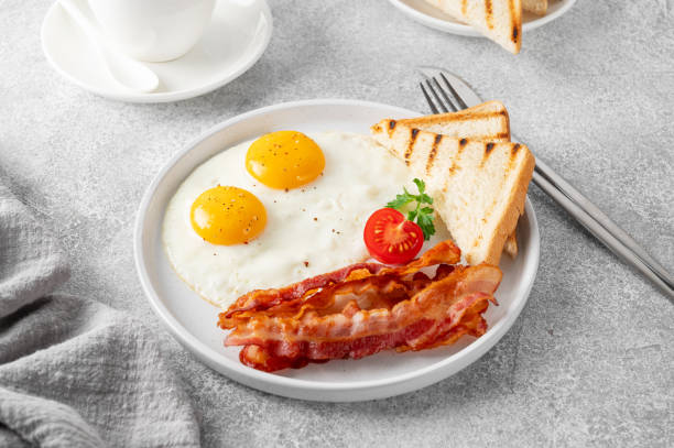 istock Breakfast with fried eggs, bacon and toasts on a white plate with cup of coffee on a stone background. Top view, selective focus. 2157554382