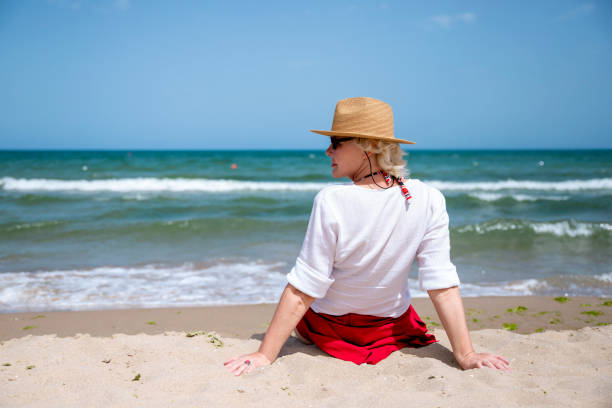 Back view of relaxed Senior Woman sitting on sand and looking at sea view from the beach. Positive lifestyle and wellbeing of 60 years old women. Summer, holidays, vacation, travel concept. Copy space