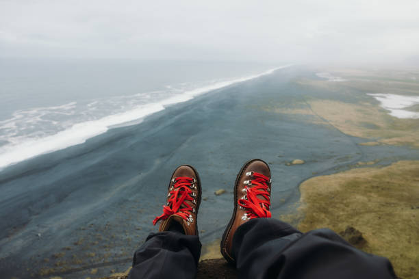 Dramatic view of women's legs wearing hiking boots with red laces sitting with a view from above of scenic endless black sand beach at Dyrhoaley, South Iceland
