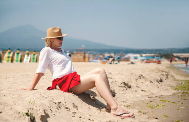 Mature Woman sitting and relaxing on the sand beach with Volcano Etna in the background in Catania, Sicily. Copy space