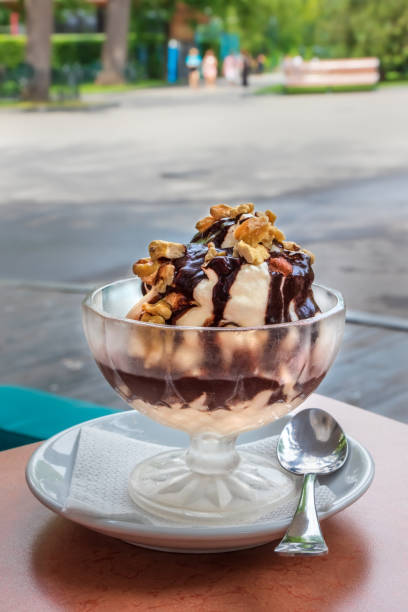 White vanilla ice cream with chocolate topping and walnuts in a glass on the table, outdoor cafe area on a sunny summer day
