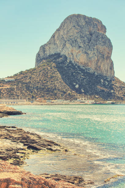 This captivating photo captures the imposing Penon de Ifach rising majestically from the Mediterranean Sea in Calpe, Spain. The towering rocky formation stands as a stark contrast against the clear blue sky, with the shimmering waters below adding to the tranquil yet awe-inspiring scene. The coastal landscape includes a quaint harbor and buildings that highlight the harmony between natural grandeur and human presence.