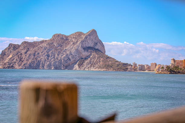 This stock photo showcases the stunning Penon de Ifach in Calpe, rising mightily against a blue sky, bordered by modern urban development and a historic watchtower. The natural framing subtly directs the viewer's gaze to the harmonious blend of nature's grandeur and human ingenuity.