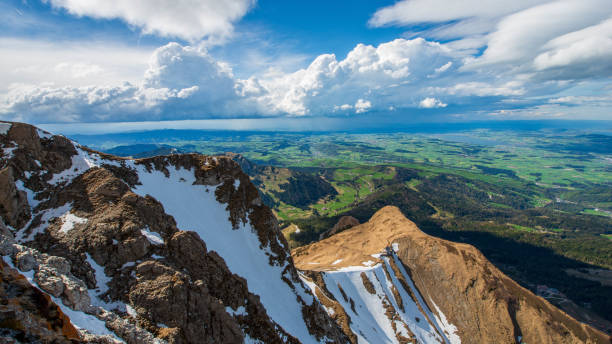 This awe-inspiring photograph captures the grandeur of towering mountain peaks partially covered in snow, juxtaposed against a vast, lush green valley below. The dramatic cloud formations in the sky add a dynamic element, creating shadows and highlights across the rugged terrain. The sharp contrast between the rocky mountains and the vibrant patchwork of fields and forests in the distance emphasizes the diverse natural beauty of the landscape. The panoramic view stretches beyond the horizon, inviting viewers to marvel at the majestic scenery and the harmony between the high-altitude wilderness and the serene lowland plains.