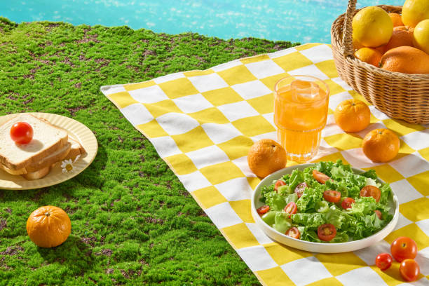 istock Beach shore grass hill scene with summertime picnic from above view, vacant space on yellow white checkered carpet for product displaying. Fresh fruit, food and beverage for picnic meal placed around 2157634528