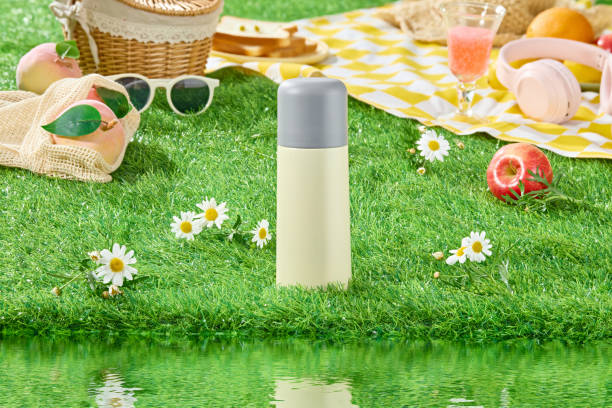 istock Picnic layout on green grass along the lake scene, from front angle. An unlabeled bottle placed in the middle, dotted with a few tana chrysanthemums, picnic tools decoration in back 2157641609
