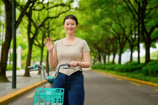 Woman Waving While Walking Bike on Green Tree-lined Path