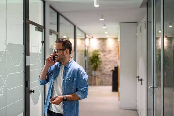 istock Happy male software designer discussing project over mobile phone while standing in office corridor 2157651023